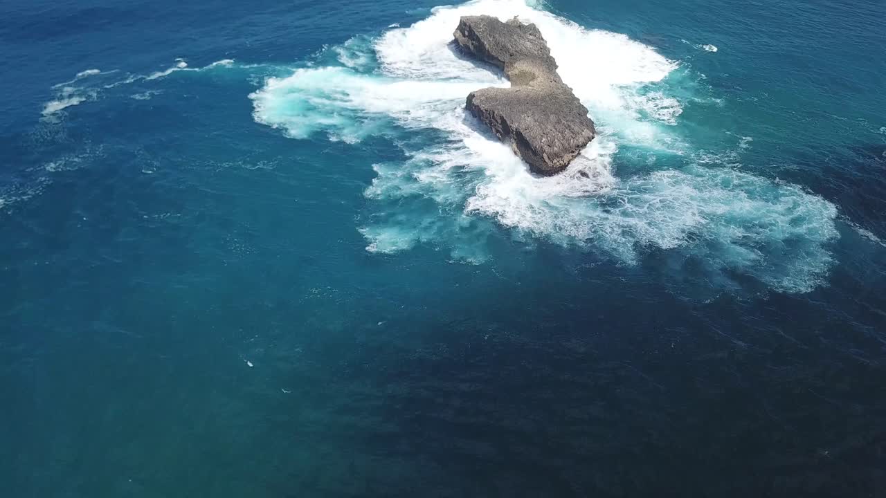una toma panorámica que revela una isla rocosa aislada siendo golpeada por olas desde todas direcciones