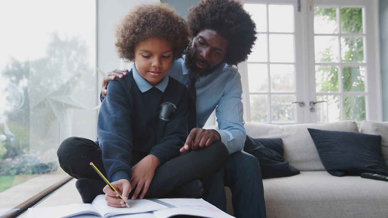 Middle aged black father sitting behind his sone helping his pre-teen son with homework in their living room at home, front view, close up