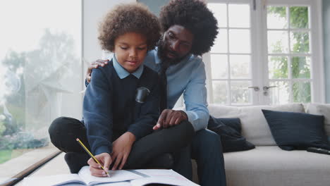 Padre-Negro-De-Mediana-Edad-Sentado-Detrás-De-Su-Hijo-Ayudando-A-Su-Hijo-Preadolescente-Con-La-Tarea-En-Su-Sala-De-Estar-En-Casa,-Vista-Frontal,-De-Cerca