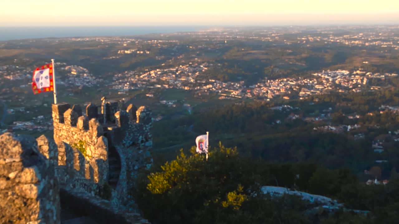 Golden hour sunset view from the Moorish Castle in Sintra Portugal showing ancient stone walls stairways, a waving castle flag overlooking hills villages and distant coastline with no people present
