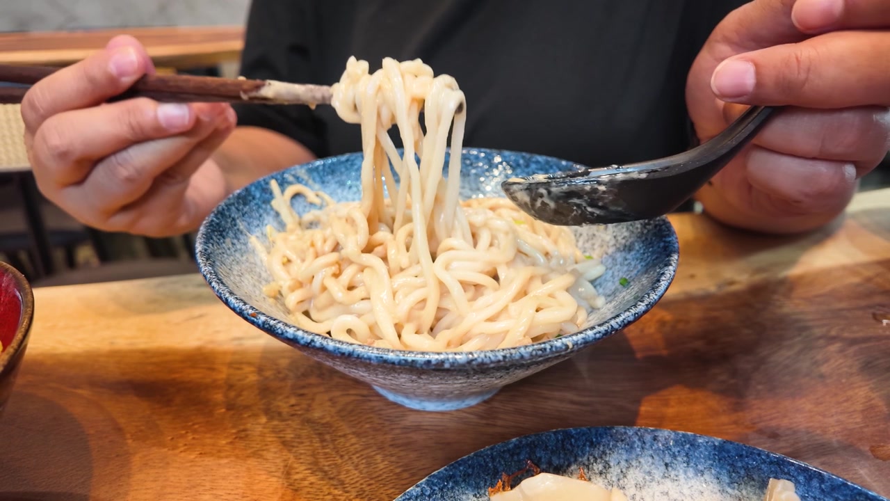 Primo piano di mani che mescolano noodles Ma Jiang Mian in una ciotola blu su legno.