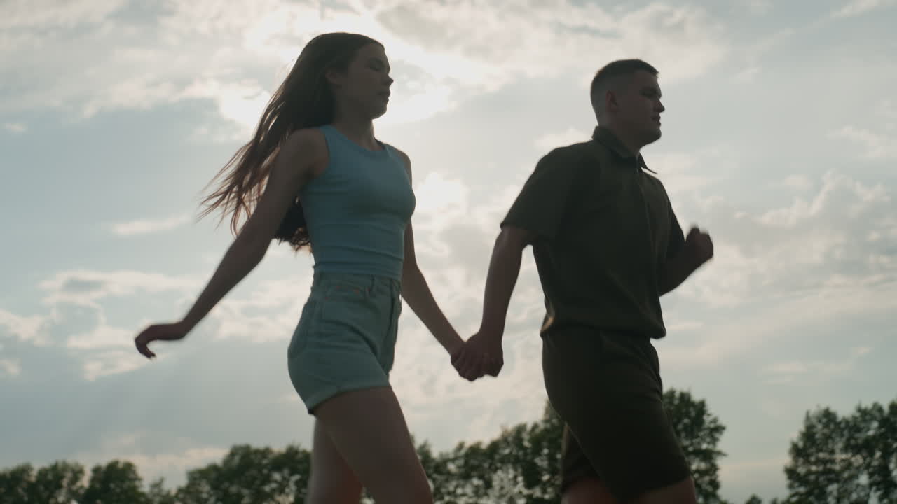 side view of couple running through open green field under bright sun flare, woman hair swaying as she gasps for breath in exhaustion while man powers stride