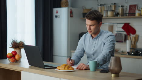 Hombre-Tomando-Notas-Trabajando-Con-Una-Computadora-Portátil-En-La-Cocina.-Freelancer-Usando-Tecnología-Inalámbrica