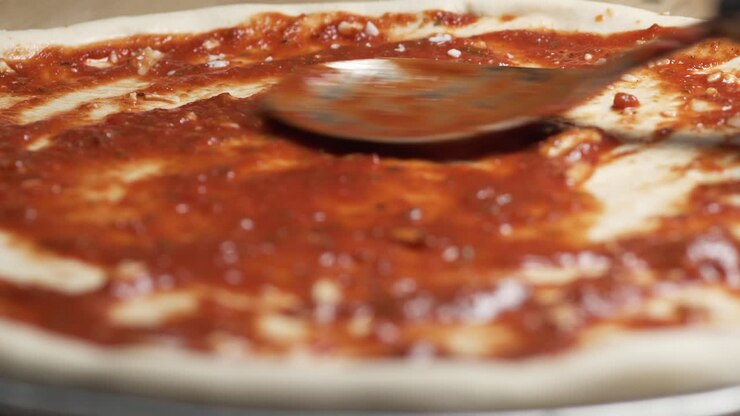 Mano di un talentuoso chef che finisce di cospargere mozzarella su un impasto per pizza con salsa rossa succosa, posizionato su un vassoio d'acciaio su un bancone di legno.