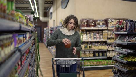 front view of caucasian woman walk with a cart near shop shelves choosing a glass jar in grocery market and put it to the cart. female customer checking product ingredients. supermarket, sale, shopping, assortment, consumerism concept