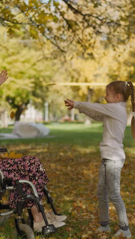 girl with long braids throws up autumn leaves heap above mother in wheelchair. couple raises hands laughing joyfully. family recreation in nature