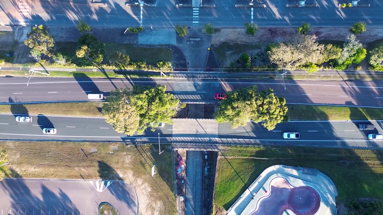 Drone aerial landscape of car vehicles travelling along a multi-lane highway road with a pedestrian overpass bridge beside a carpark and an urban skatepark in Bateau Bay Central Coast NSW Australia