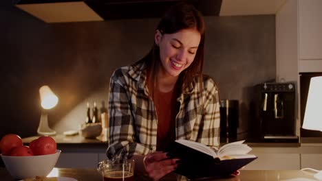 Happy-brunette-girl-in-a-plaid-shirt-and-an-orange-T-shirt-stands-near-the-table-in-the-kitchen-and-reads-her-book-while-spending-leisure-time-and-relaxing-in-a-modern-apartment-in-the-evening