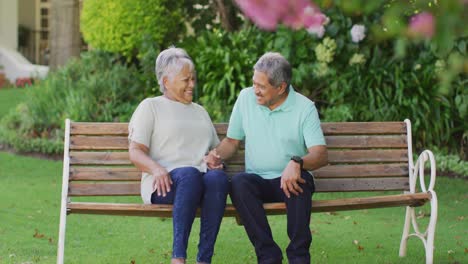 video of happy biracial senior couple embracing and sitting on bench in garden