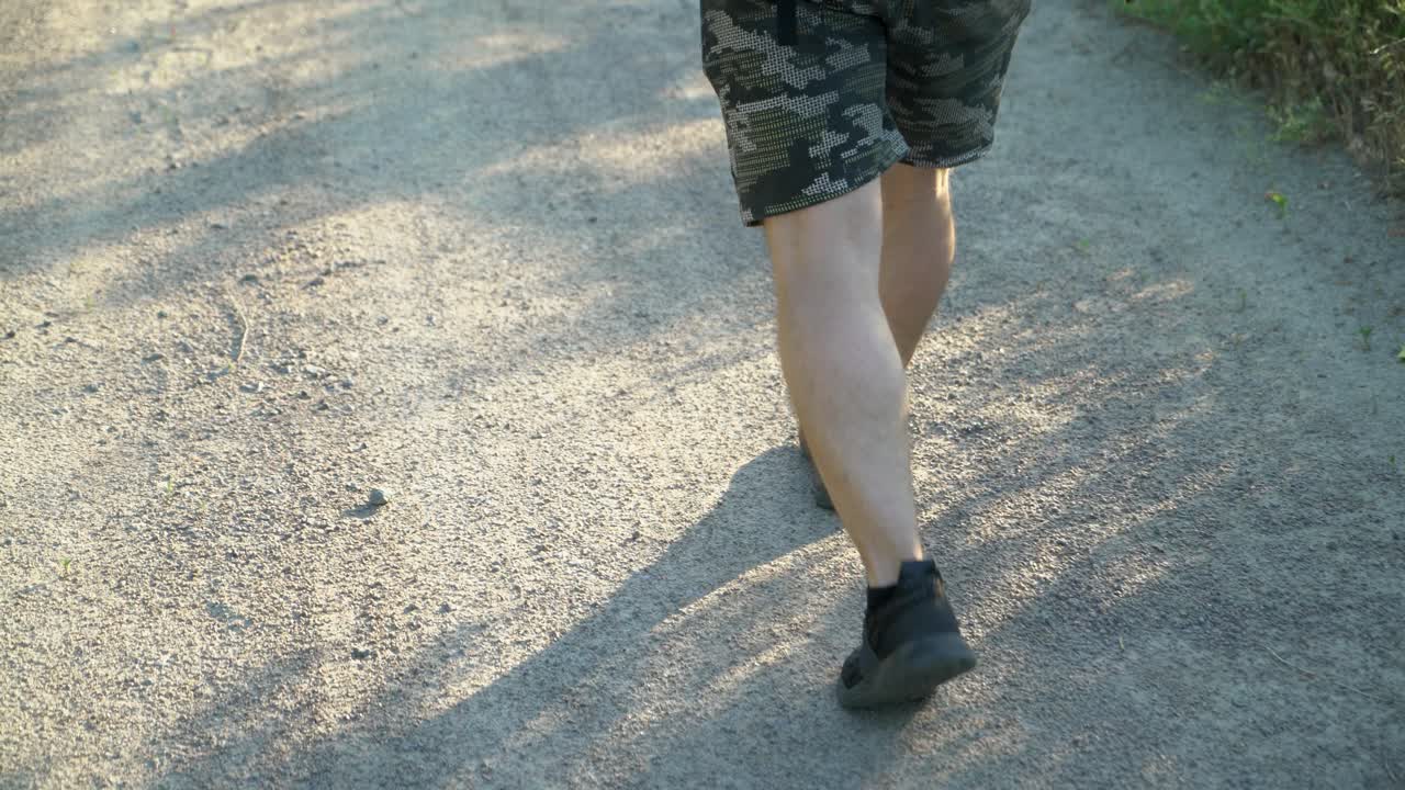 A man in shorts and black sneakers is walking on a dirt road on a summer day. A tourist goes hiking.