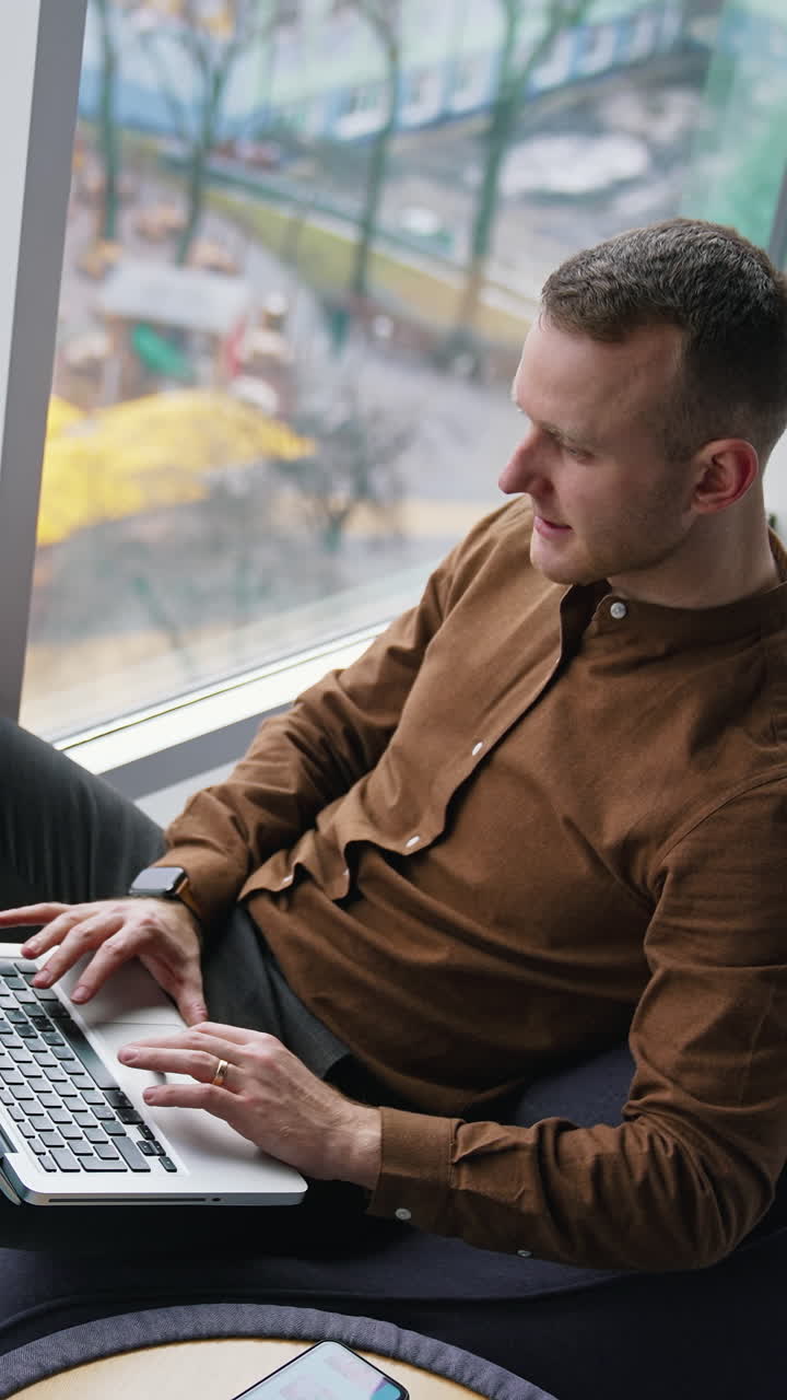 Handsome employee with a laptop in office. Young businessman sitting in a comfortable chair and works near the window with city landscape. Top view. Vertical video