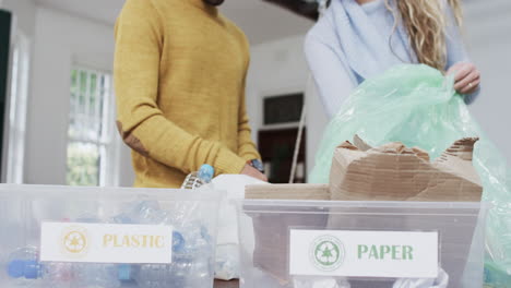 Midsection-of-diverse-couple-segregating-plastic-bottles-and-paper-at-home,-slow-motion