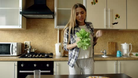 cheerful young funny woman dancing and singing with lettuce microphone while cooking breakfast