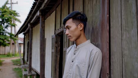 young southeast asian man standing near wooden house in rural village, slowly turning head and gazing directly at camera with contemplative expression