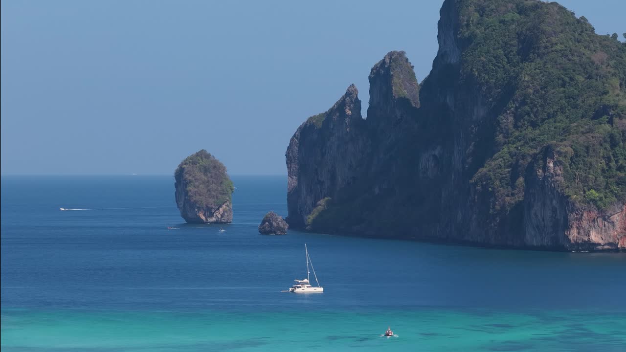 Panoramica di una barca a vela bianca ancorata in acque calme e turchesi accanto a scogliere calcaree tropicali.