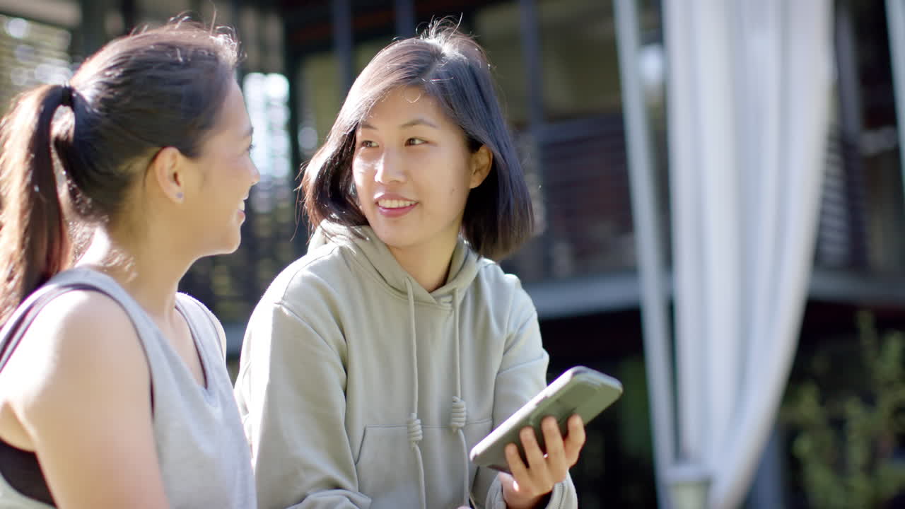 Happy asian female friends with water bottle and smartphone laughing on terrace, slow motion