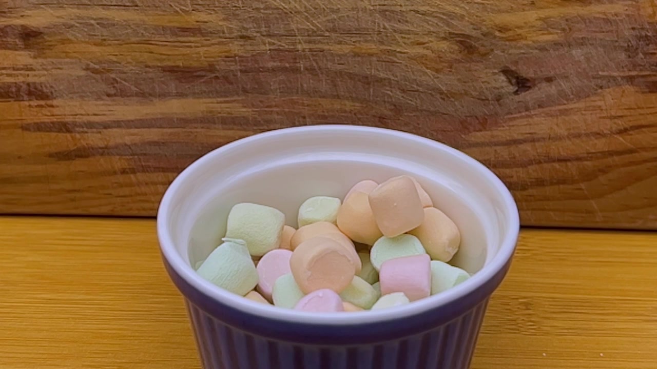Nærbillede af farverige pastel-marshmallows arrangeret i en hvid skål på et træbord, der viser blød tekstur og assorterede lyserøde, gule, grønne og hvide stykker