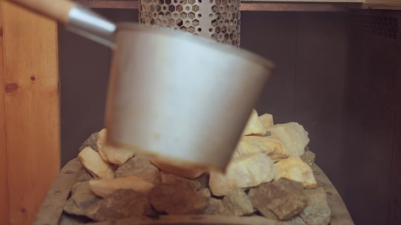 close up of person holding metal ladle pouring water onto hot sauna stones with steam rising, capturing soothing atmosphere of wellness ritual in wooden room