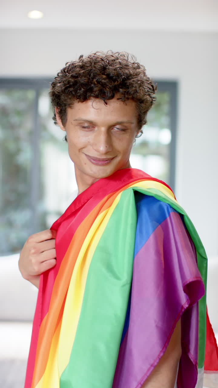 Vertical video of portrait of happy biracial man holding rainbow lgbt flag at home, slow motion