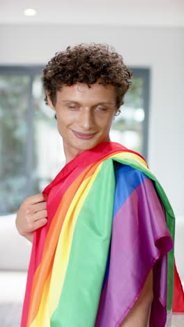 vertical video of portrait of happy biracial man holding rainbow lgbt flag at home, slow motion