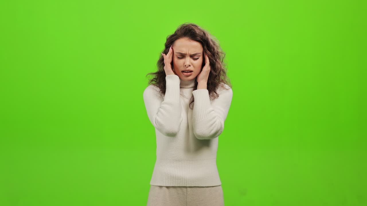The woman is standing and looking at the camera. She is holding her head and has a headache. She is standing on a green background. Green screen. 4K.