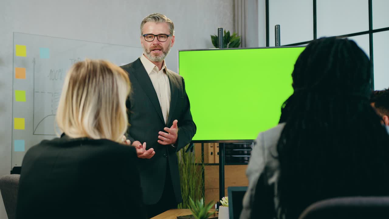 Business concept where likable confident experienced bearded 50-aged businessman explaining presentation on green screen for attantive successful mixed race team of colleagues