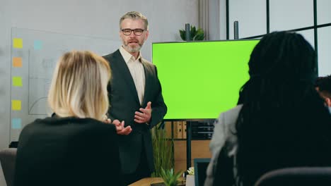 business concept where likable confident experienced bearded 50-aged businessman explaining presentation on green screen for attantive successful mixed race team of colleagues