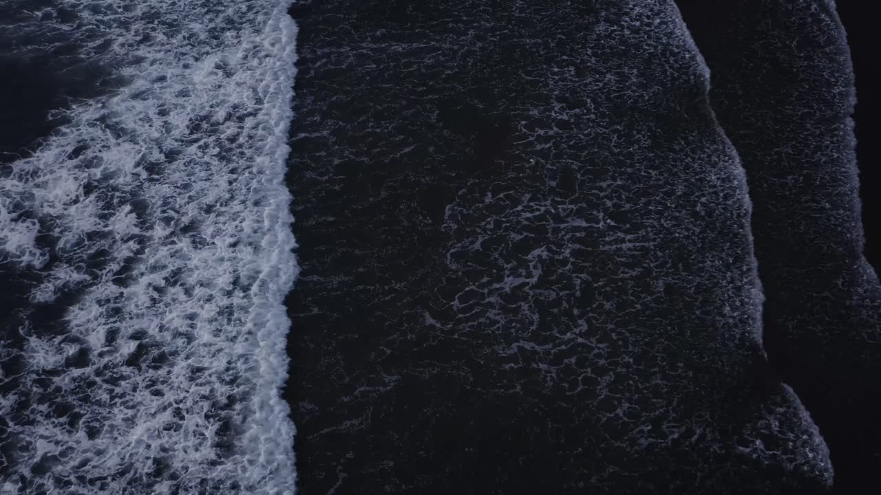 Powerful waves crash on the shore forming symmetrical foam patters. Monochrome drone footage of waves at night, Oregon Coast. Top-down aerial view