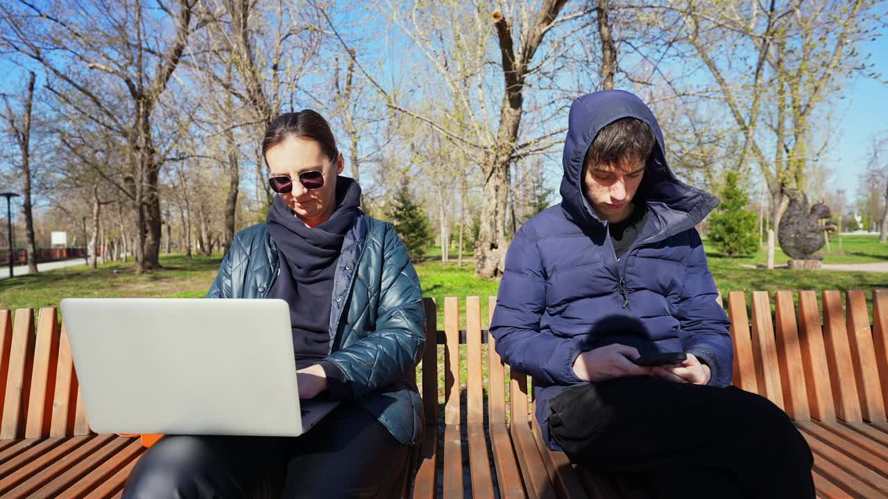 Kvinde arbejder på bærbar computer og mand bruger smartphone på parkbænk