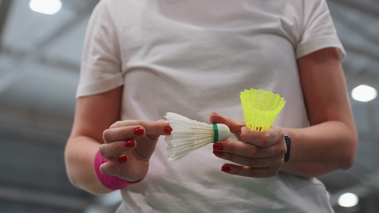 Donna caucasica che tiene i volani al chiuso, primo piano delle mani che sistemano volani bianchi e neon, polsino rosa, smalto rosso, smartwatch, camicia bianca, luci dell'arena dall'alto, concentrata prima della partita