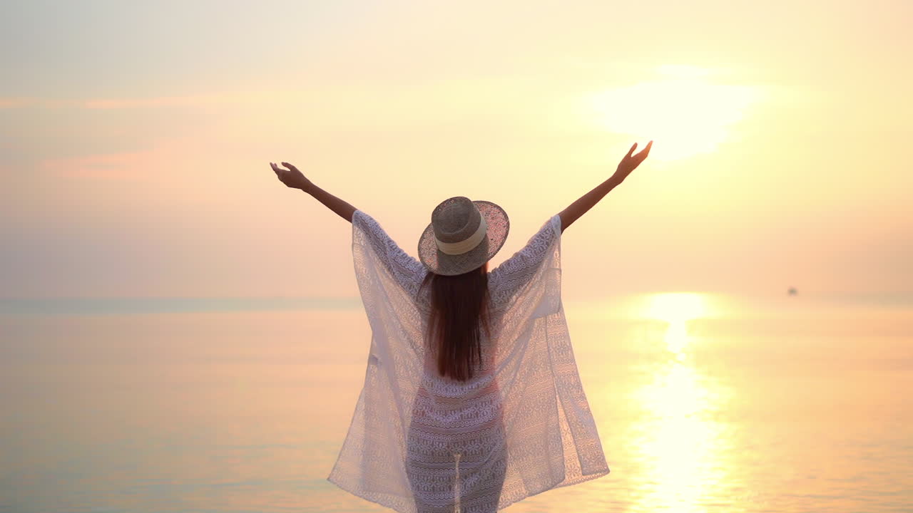 una mujer joven de espaldas a la cámara camina hacia el sol poniente levantando los brazos con alegría