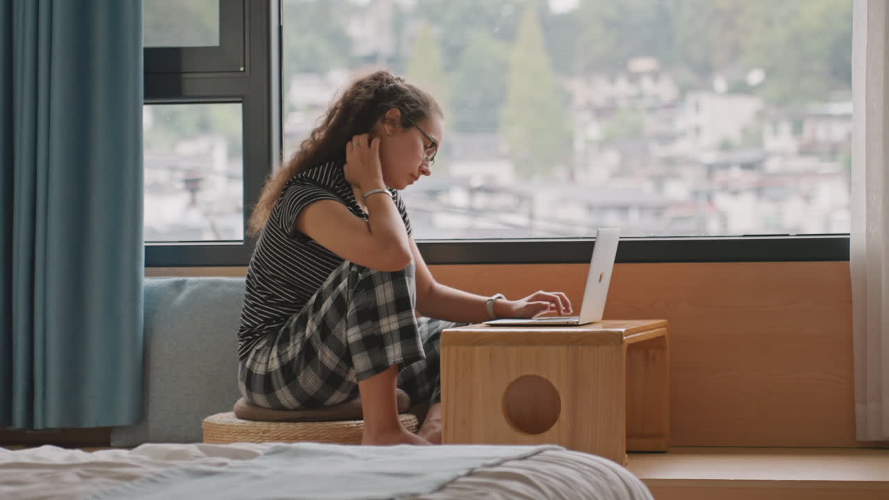 Uformel læringssituation. Studerende studerer afslappet ved vinduet. Afslappet studerende fokuseret på bærbar computer, siddende ved vinduet. Afslappet studerende arbejder flittigt på bærbar computer ved vinduet i studietiden.