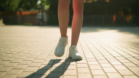 leg view of individual wearing white canvas shoes walking along interlocked path with shadow cast on ground, with partial view of roller skate, blurred background featuring greenery