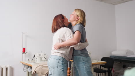 creative garment designers hug tightly inside sewing studio, playful hands celebrating successful project, denim jeans brushing, sewing machines and folded fabrics surrounding scene, joyful energy