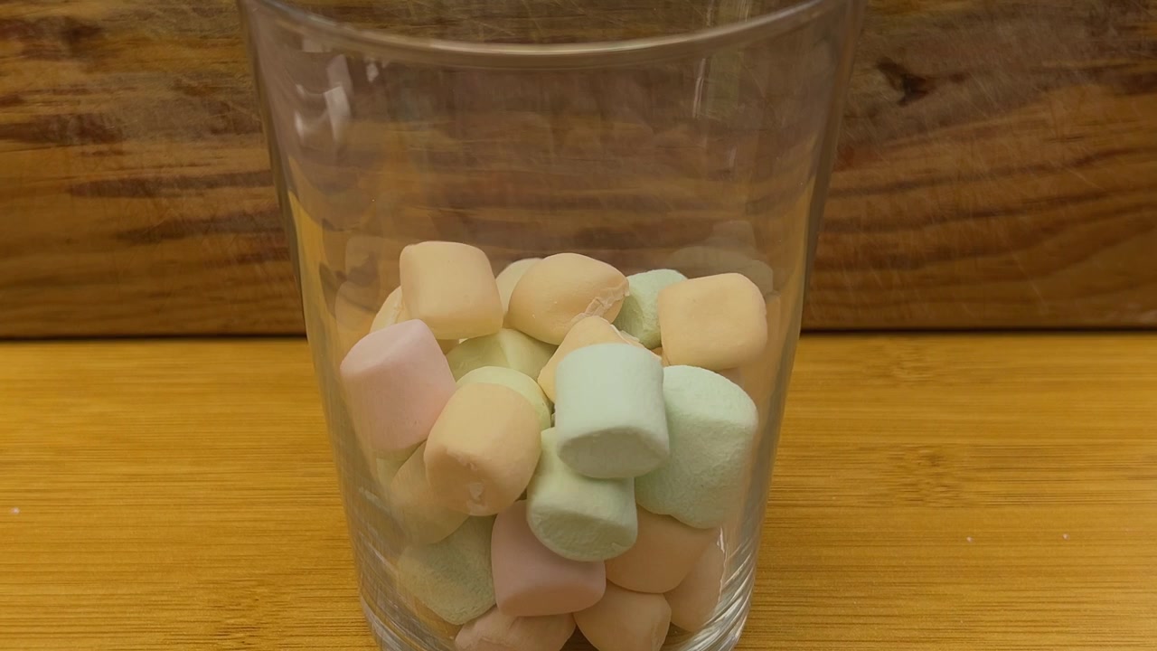 Farverige skumfiduser fylder et klart glas på et træbord, med bløde pastelkonfektstykker der falder ned i en gennemsigtig beholder i nærbillede.