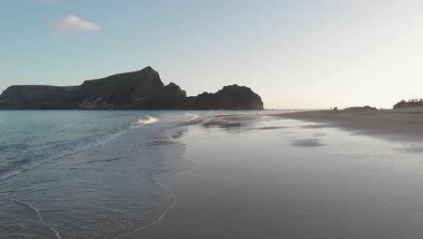 Exhilarating-flight-above-wet-sandy-beach-with-ocean-sea-waves-breaking-on-shoreline-coast-toward-silhouette-Cal-Islet-brown-island-and-mountain-peak-at-sunset,-Portugal,-aerial-approach