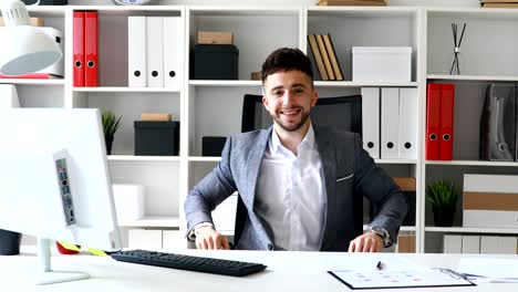 joven empresario sentado en la mesa en la oficina blanca, sonriendo y mostrando el gesto de la mano de aprobación