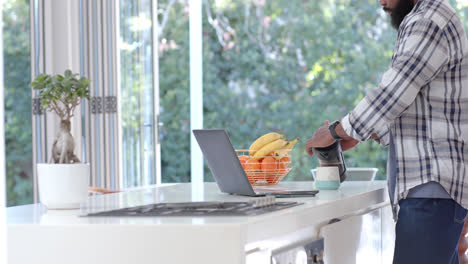 african american man making coffee and using laptop in sunny kitchen, slow motion