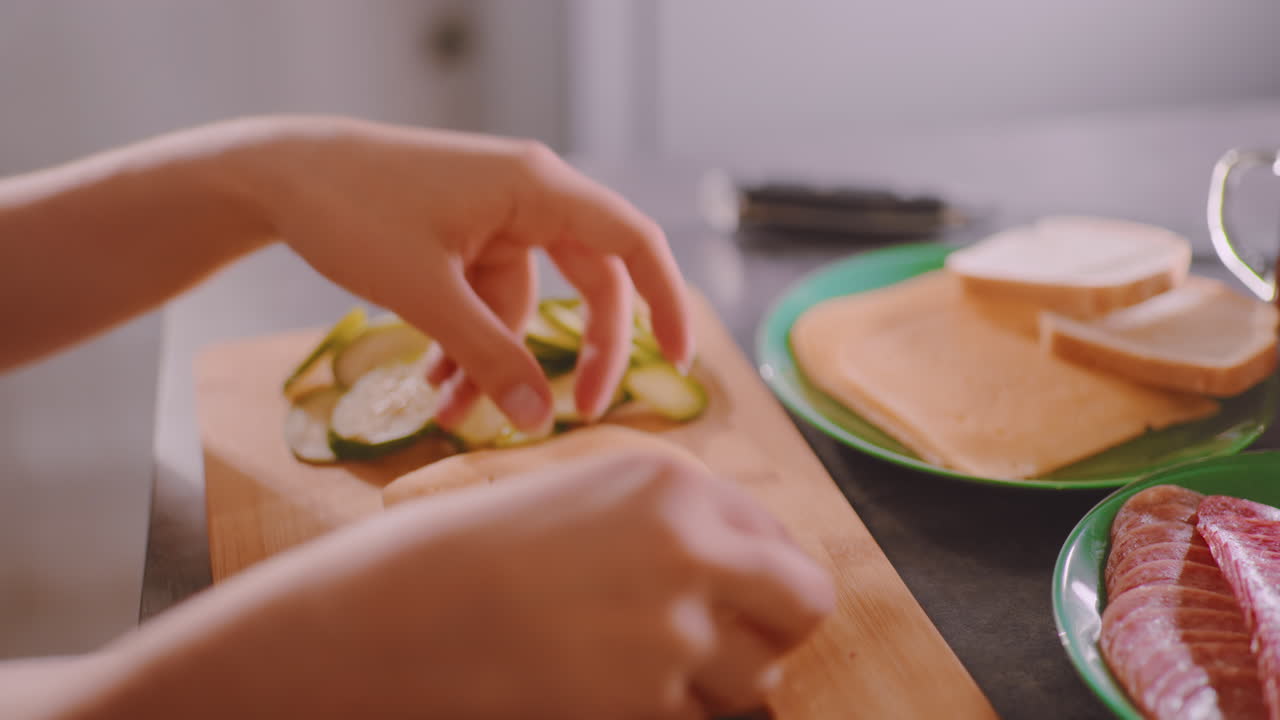 Close up of chef preparing sandwich with slices of bread, cheese, and sausage on green plate, arranging ingredients for breakfast beside cup of coffee in bright kitchen