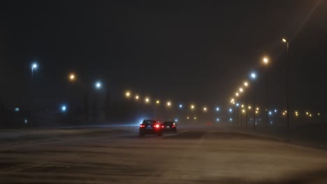 toma estática de autos conduciendo en una carretera bien iluminada durante una ventisca con nieve suelta en la carretera arrastrada por fuertes vientos por la noche