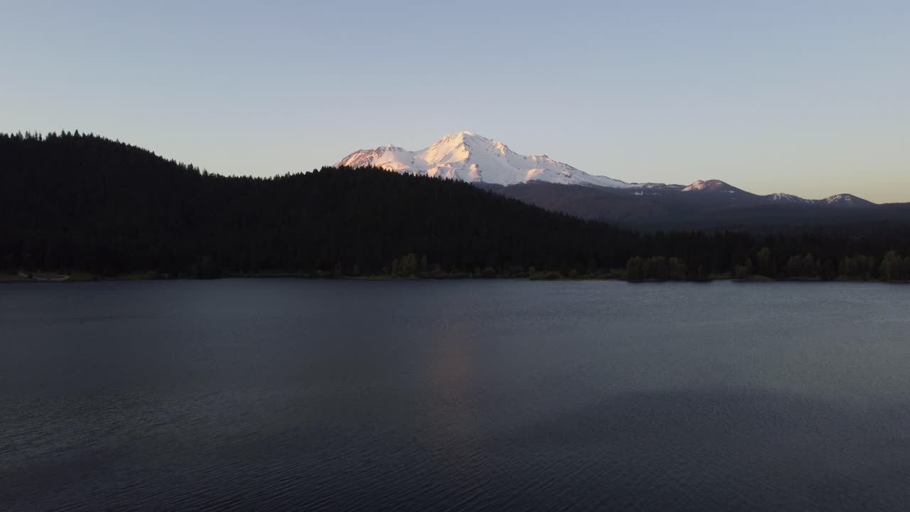 Mount Shasta volcano revealed from behind the hills at dusk over Lake Siskiyou in Northern California. Snow-covered mountain reflecting on the water as the sun sets. 4K cinematic drone shot.