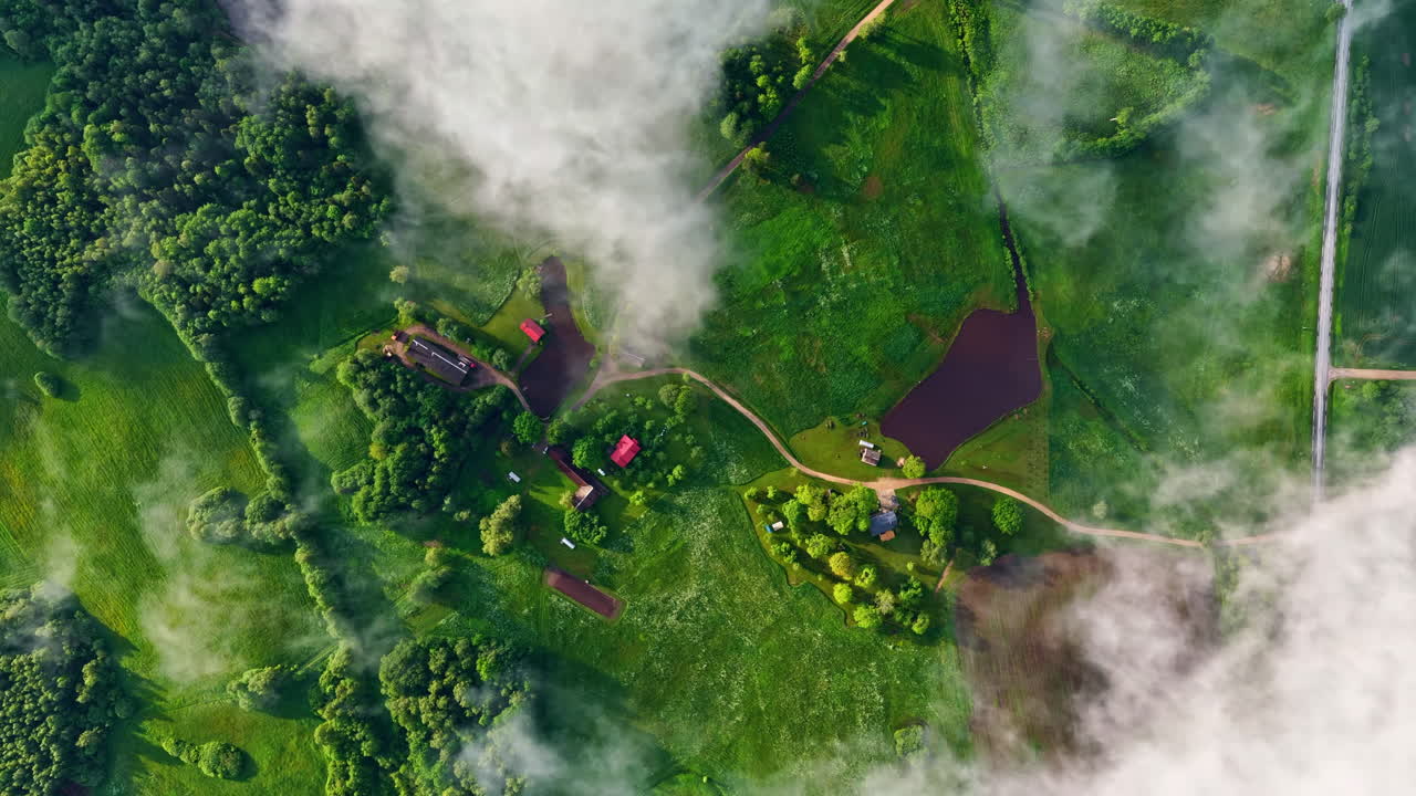 A breathtaking top-down perspective of the lush green Latvian landscape with wispy clouds or mist gently flowing through fields, forests, and rural farm buildings