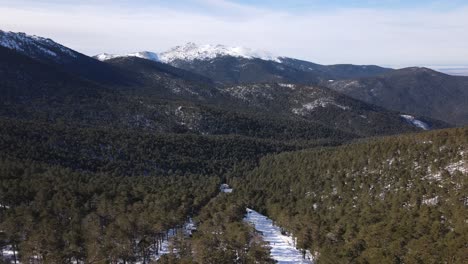 Forward-dolly-aerial-view-over-the-forest-and-ski-slope-heading-towards-snow-peaked-mountains