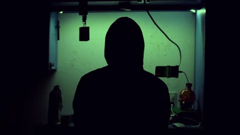 silhouette eines mysteriösen hackers, der einen hoodie trägt und in einem dunklen raum mit seinem computer und seinem telefon voller grüner programmiercodes spielt