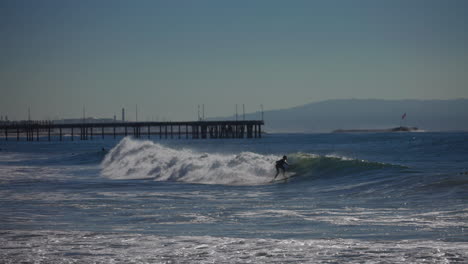 a surfer rides a wave down the line with a pier and mountains in the background