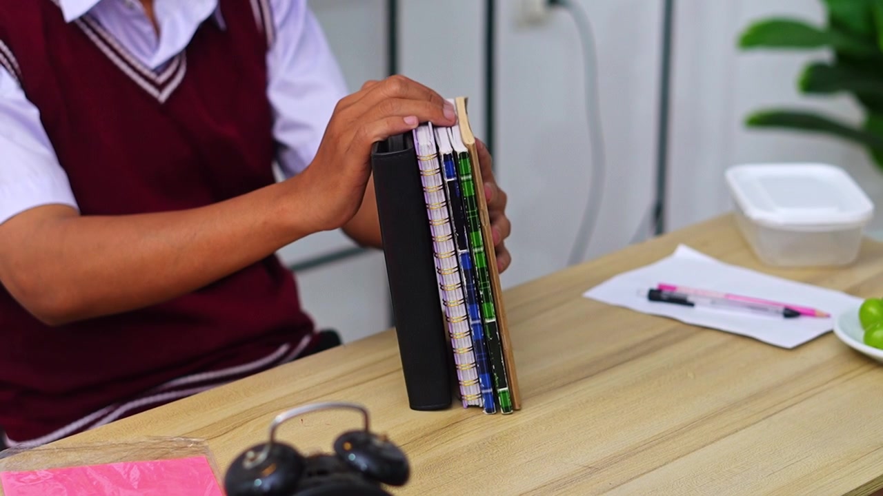Primo piano di uno studente in uniforme scolastica che organizza una pila di quaderni e agende su una scrivania di legno.