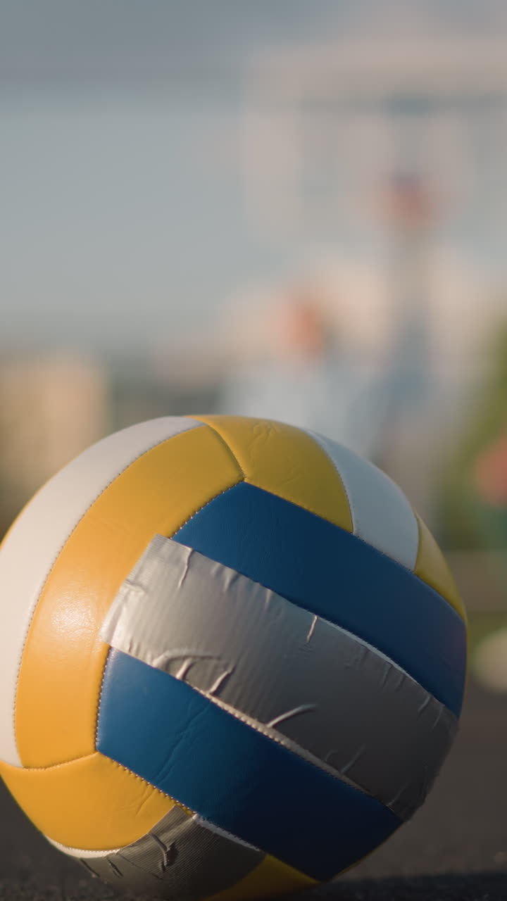 primer plano de una pelota de voleibol colocada en el suelo con fondo borroso que muestra a personas haciendo ejercicio y estirándose en un campo deportivo al aire libre con una actividad atlética vibrante