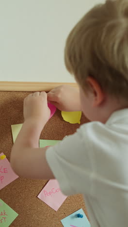 toddler attaches bright pink leaf from colored paper. child learns to work with paper playing with cork board slow motion. development of kid skills backside view closeup