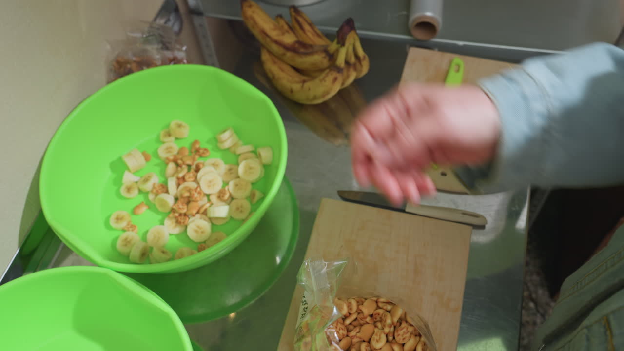 Tangan menambahkan granola ke mangkuk pisang, blogger makanan merangkai sarapan renyah dengan sereal dan buah berlapis, gerakan close-up di atas mangkuk plastik hijau di meja dapur rumah dalam rutinitas pagi yang santai