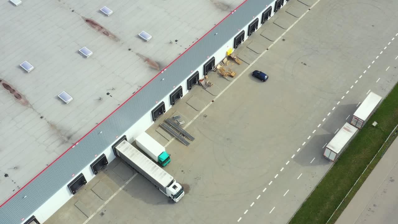 Aerial following shot of semi-trailer trucks travelling along a warehouse on the parking lot in the logistics park with loading hub.